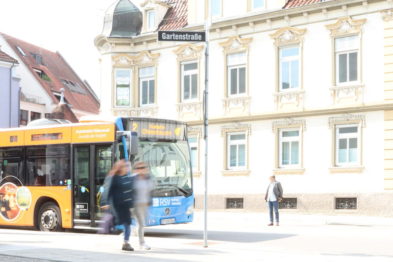 Ein Bus steht an der Haltestelle. Es sind unscharf Mesnchen zu sehen, die gerade ausgestiegen sind.
